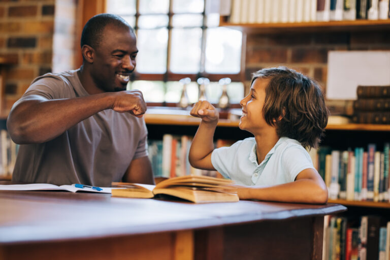 Teacher and student giving each other a high five in a library. Elementary school boy having a private lesson with his teacher. Tutoring and student support in a primary school.